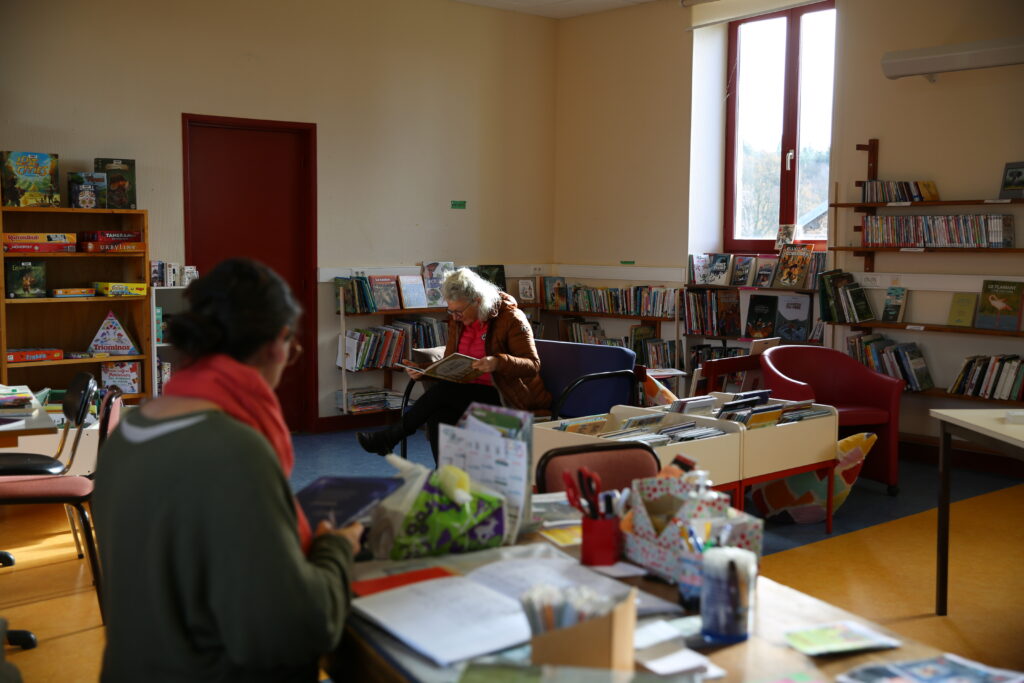Une bibliothécaire bénévole, de dos enregistre un document à son bureau. Au fond on voit une lectrice consulter un livre sur un canapé, dans le coin jeunesse, elle sourit.
